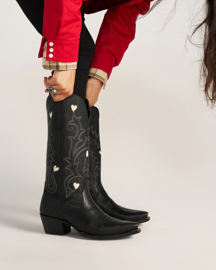 The Annie in Midnight Cowhide. Person in a red shirt and black pants bends down to adjust tall black Midnight cowboy boots with white heart details and decorative stitching.