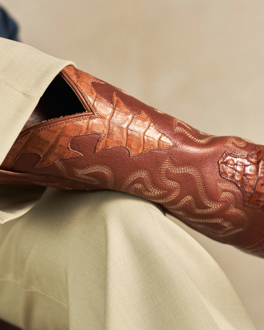 The Garrett in Pecan Hornback Caiman. Close-up of a brown leather Hornback Caiman cowboy boot with decorative stitching, resting on a crossed knee in light-colored pants.