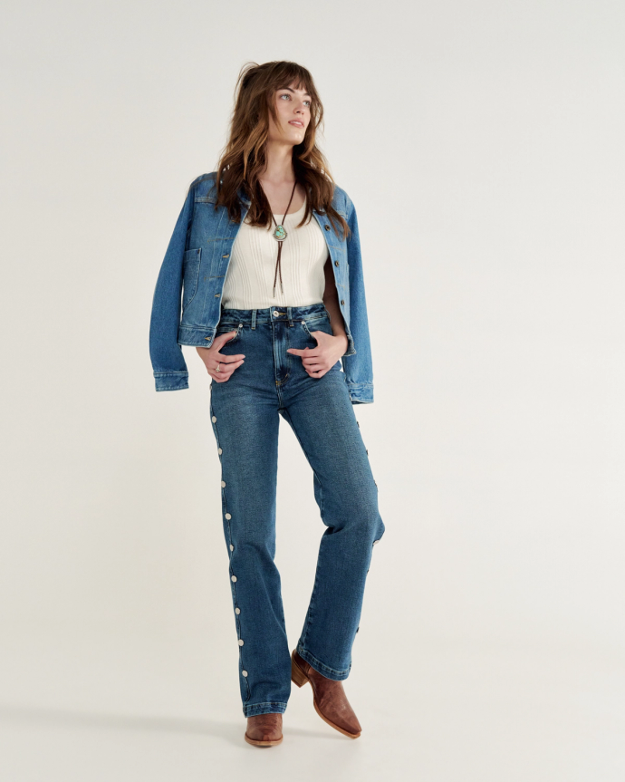 Woman wearing a denim jacket, white top, buttoned jeans, brown boots, and a bolo tie, stands posing against a plain background.