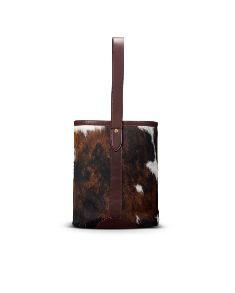 A cylindrical bucket bag with brown and white cowhide exterior, a single brown leather strap, and leather trim, displayed against a plain white background.