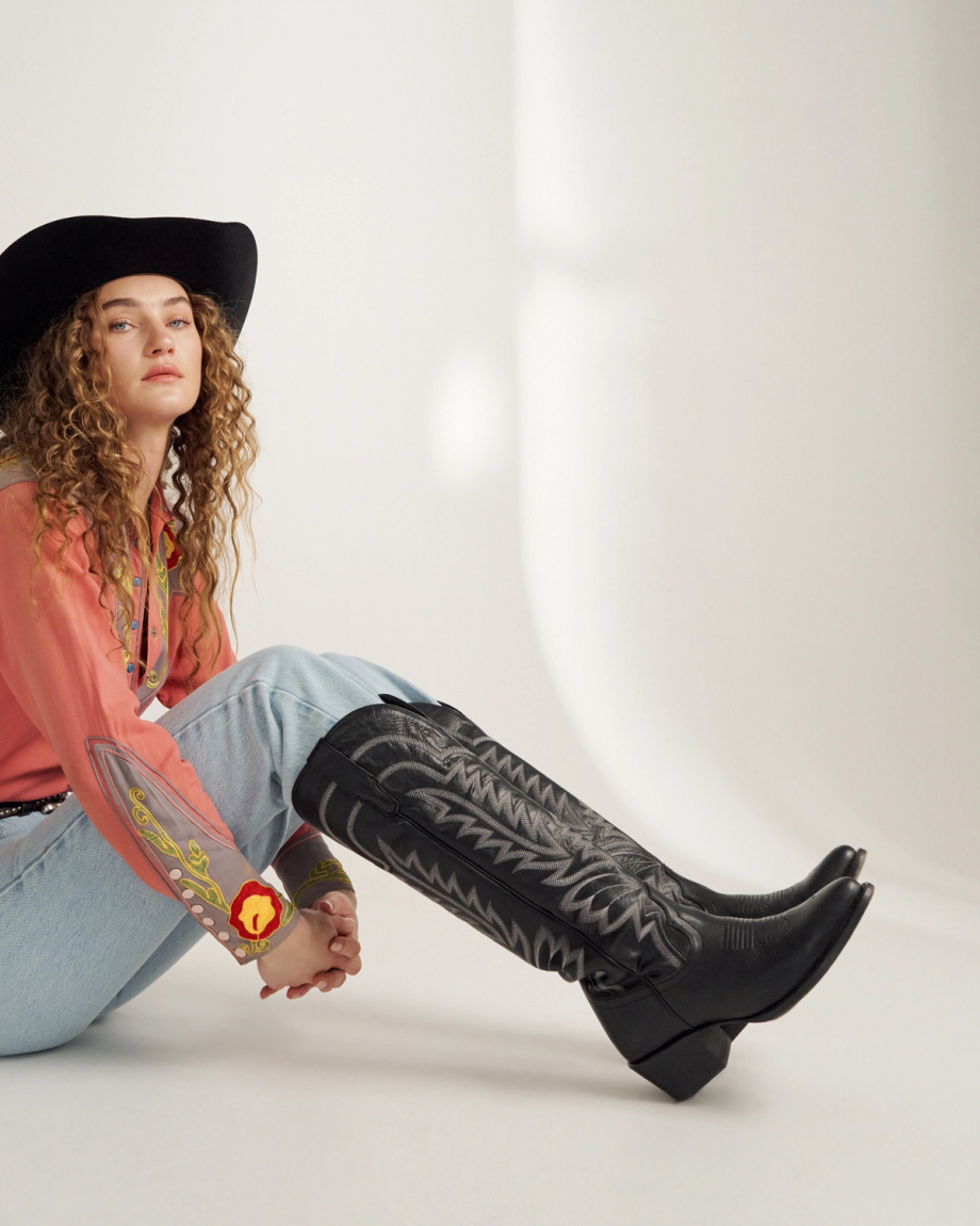 A person in a black cowboy hat, coral western shirt, light blue jeans, and black cowboy boots sits on the floor against a white background.
