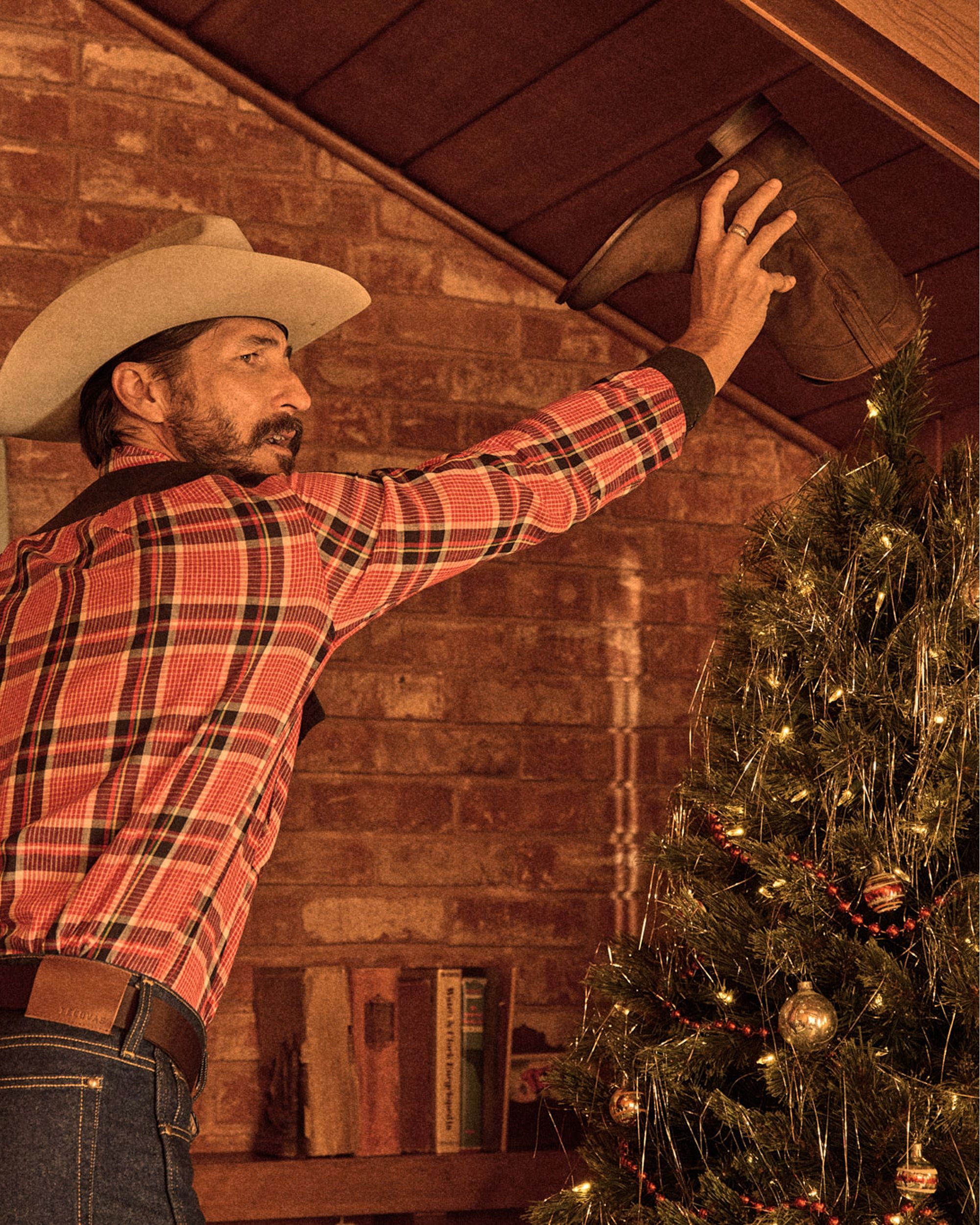 A man in a plaid shirt and cowboy hat places a brown cowboy boot atop a decorated Christmas tree in a rustic room.