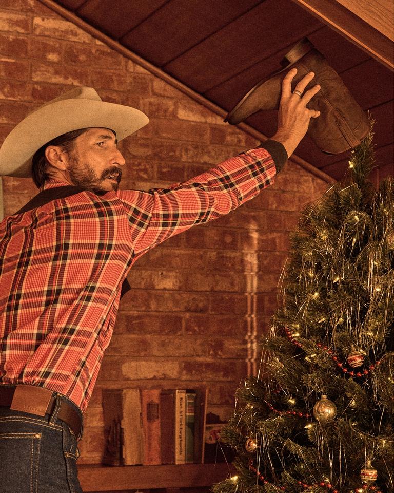 A man in a plaid shirt and cowboy hat places a brown cowboy boot atop a decorated Christmas tree in a rustic room.
