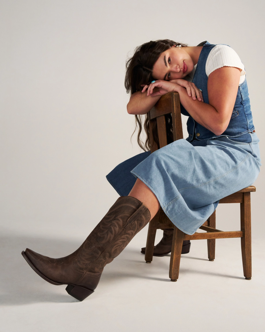 A woman in a denim dress and brown The Annie Wide Calf Goat cowboy boots sits sideways on a wooden chair, resting her arms and head on the chair’s backrest, looking at the camera as if waiting in a cozy cafe.