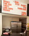 A food counter with a menu board listing items such as brisket, pork ribs, turkey, lamb chops, sausage, green beans, spaghetti, stew, charro beans, pudding, and cobbler. Two refrigerators are behind the counter.