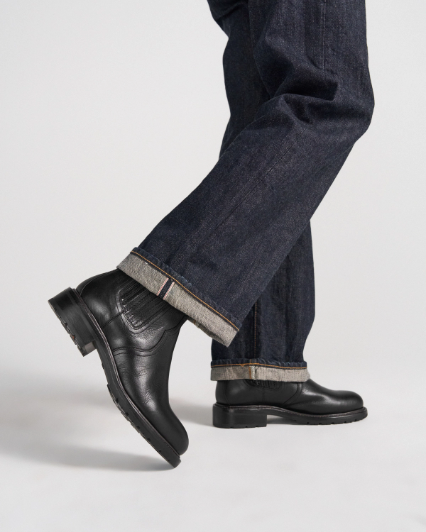 The Chelsea in Midnight Cowhide. Person wearing dark cuffed jeans and black leather boots, standing with one foot slightly raised against a plain background—the Midnight Cowhide boots add a refined edge to this timeless look.