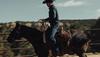A person in a cowboy hat rides a horse across a field with a wooden fence and snow-capped mountains in the background.