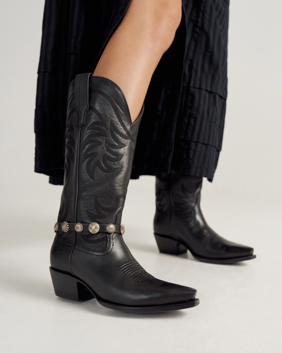 A person wearing a black skirt and black cowboy boots with decorative silver conchos stands on a light background.