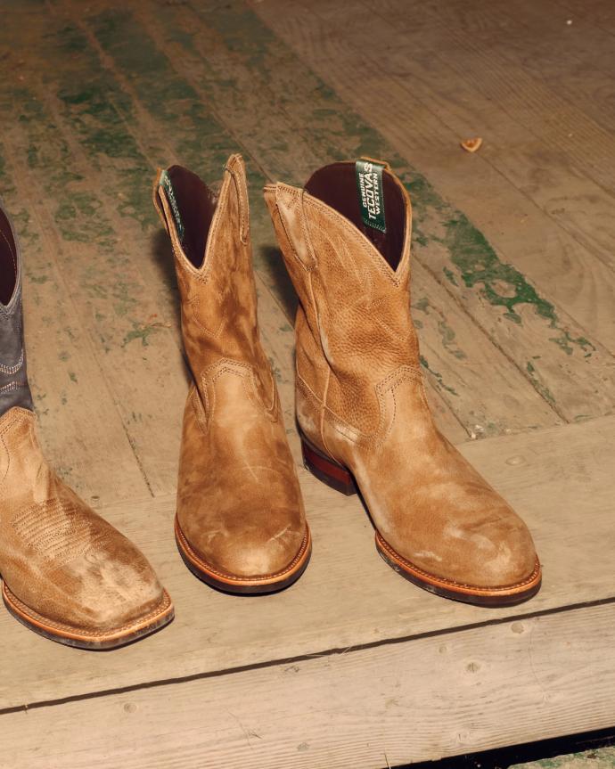 Two pairs of cowboy boots, one tan and one brown with black tops, sit on a worn wooden floor.