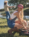 A woman wearing denim overalls, a white tank top, and a red cap kneels by a tackle box, while a man in a cowboy hat and jeans casts a fishing rod by a pond in the background.