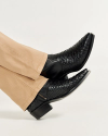 A person wearing light beige pants and black textured leather boots rests their feet against a white background.
