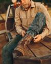 A man sitting on the back of a truck adjusts his camouflage-patterned boot, wearing a beige shirt and blue jeans.