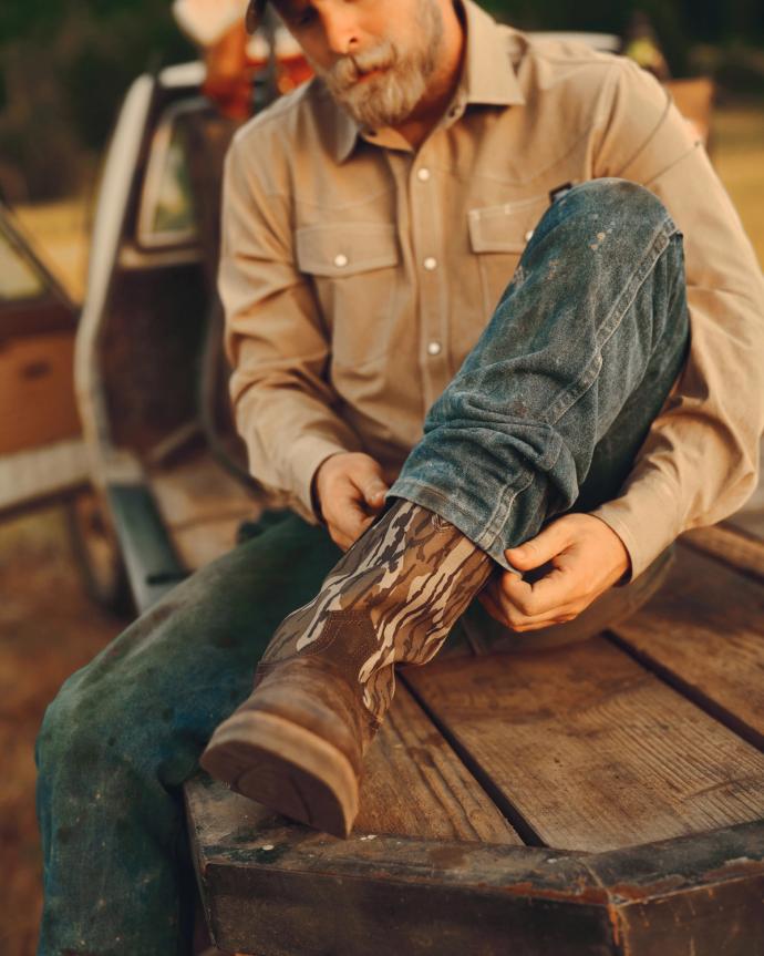 A man sitting on the back of a truck adjusts his camouflage-patterned boot, wearing a beige shirt and blue jeans.
