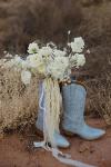 A bouquet of white flowers perched atop a pair of light blue cowboy boots on a sandy terrain.
