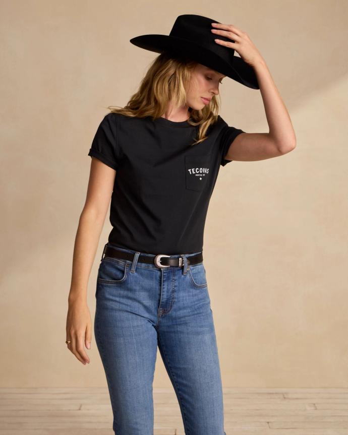 A person in a black T-shirt and cowboy hat adjusts their hat. They are wearing blue jeans and standing against a neutral background.