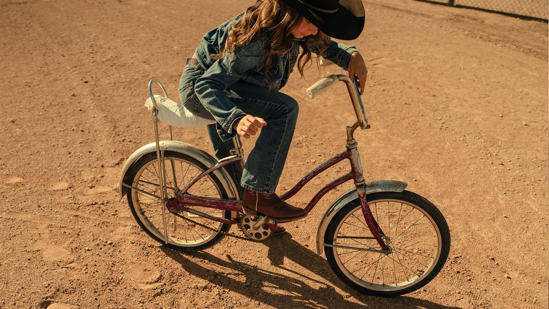 A child wearing a cowboy hat and denim outfit sits on a vintage bicycle on a dirt ground.