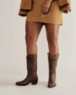 Person wearing a tan suede skirt and jacket, with brown cowboy boots, standing against a plain light background.