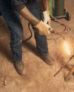 The Rugged Round Toe in Umber / Sage Cowhide. A person in The Rugged Round Toe Cowhide boots, jeans, and gloves uses a blowtorch to heat a metal rod on a sandy surface.