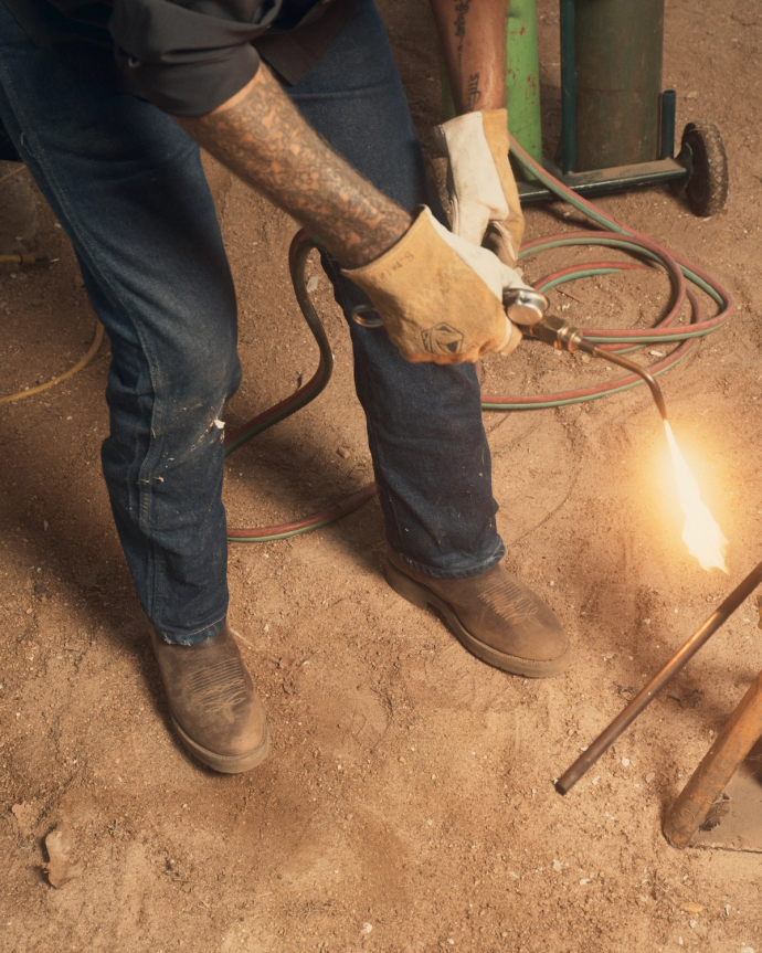 The Rugged Round Toe in Umber / Sage Cowhide. A person in The Rugged Round Toe Cowhide boots, jeans, and gloves uses a blowtorch to heat a metal rod on a sandy surface.