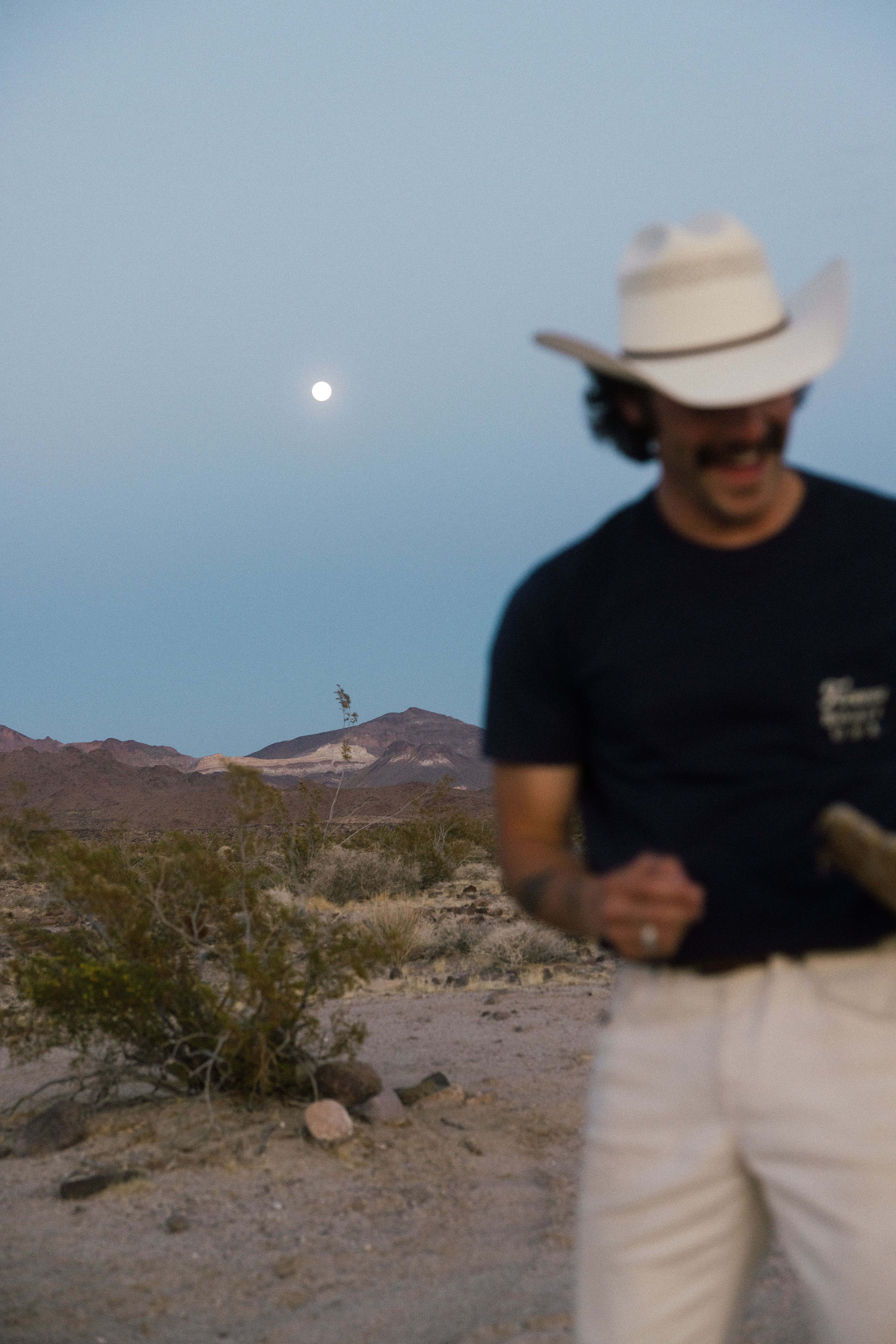 A man in a cowboy hat and sunglasses stands in a desert landscape at dusk, with mountains and a full moon visible in the background.