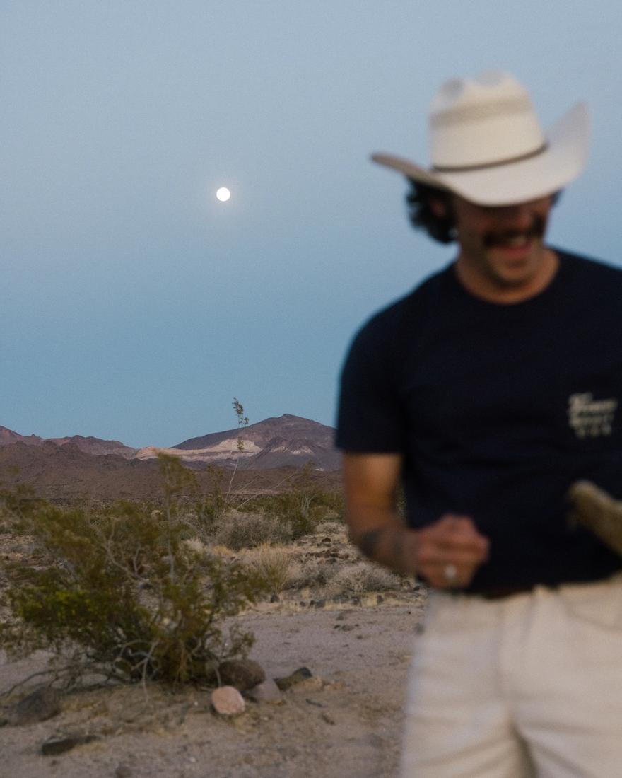 A man in a cowboy hat and sunglasses stands in a desert landscape at dusk, with mountains and a full moon visible in the background.