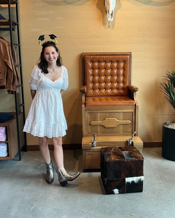 A woman in a white dress and metallic boots stands next to a leather chair with footrests. She wears a headband with small graduation caps. The room has a beige wall and various furnishings.