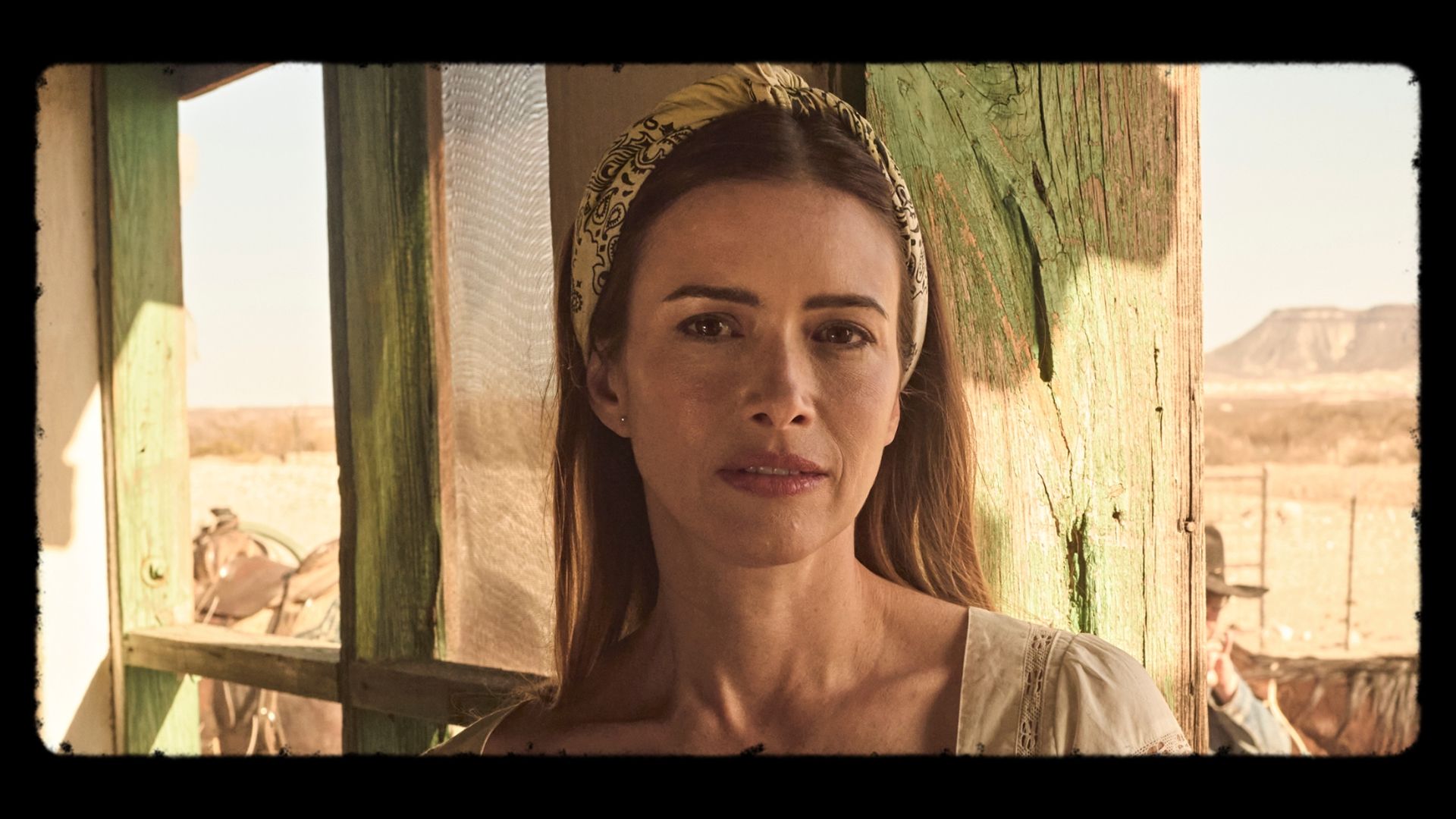 A woman with long brown hair and a yellow headband stands in front of a weathered green wooden door in a sunlit, rural setting.