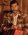 A man with a beard wearing a plaid coat sits indoors, holding a red gift box with a festive design.