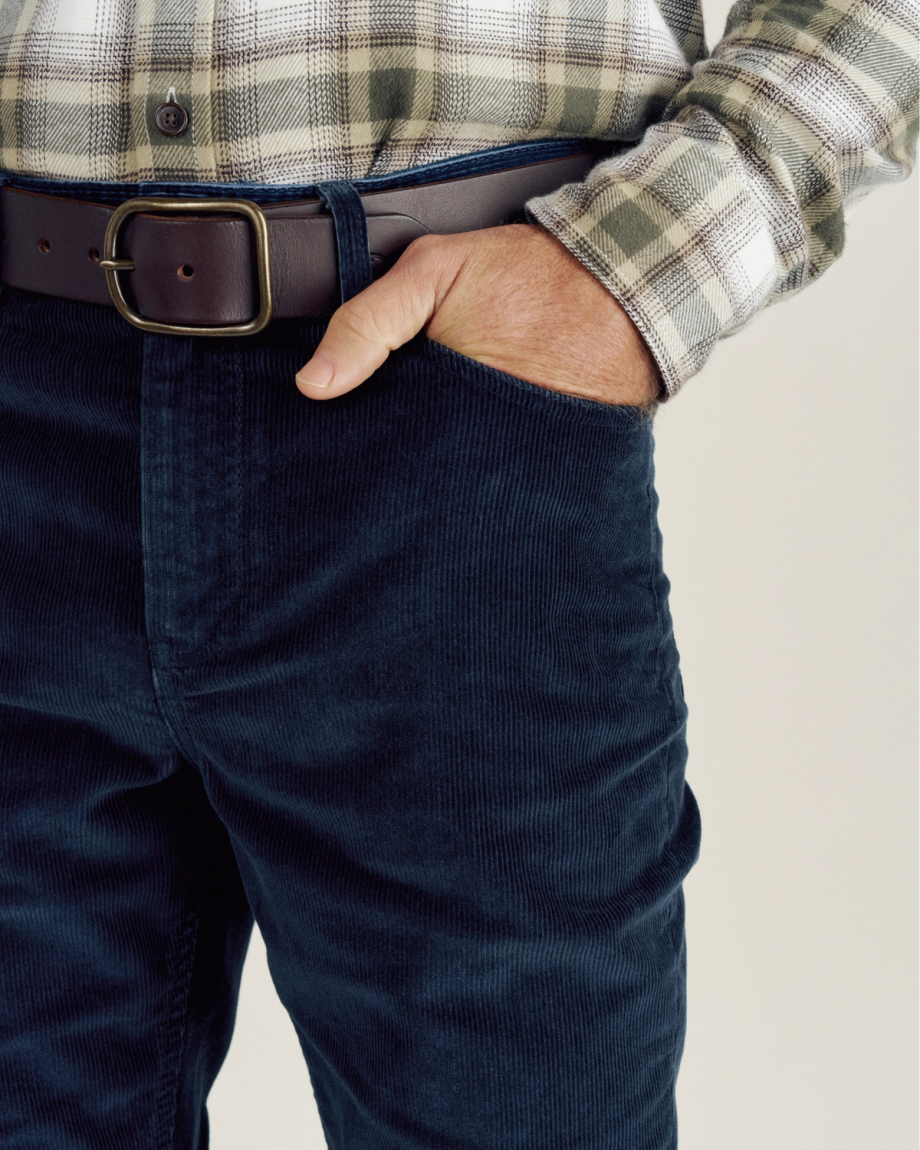 A person wearing a plaid shirt, dark belt, and navy corduroy pants with their right hand in the front pocket.