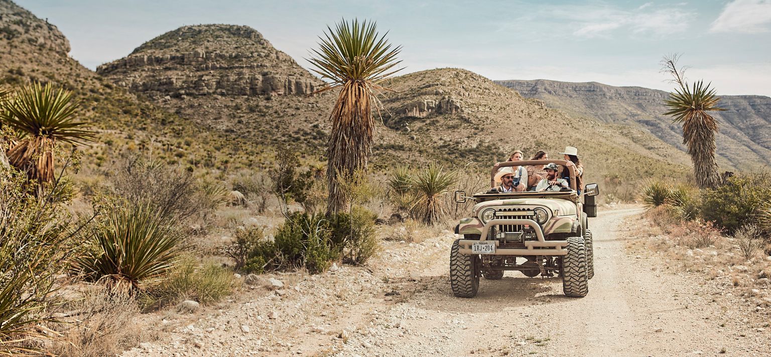 A group of people enjoying a ride in a vintage jeep on a dusty desert road, surrounded by cacti and rugged mountains.