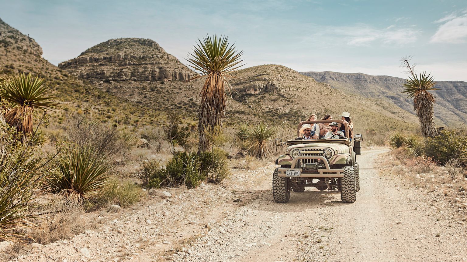 A group of people enjoying a ride in a vintage jeep on a dusty desert road, surrounded by cacti and rugged mountains.