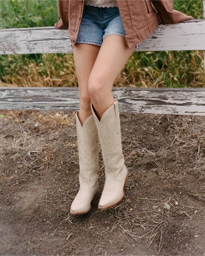 Person wearing a brown jacket, denim shorts, and cream-colored cowboy boots sits on a white wooden fence outdoors.
