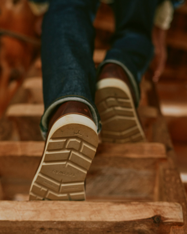 The Knox Lace Up in Cognac Cowhide. Person wearing dark blue jeans and cognac cowhide boots with white lugged soles climbing a wooden ladder.