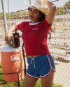 Woman in a red shirt and blue shorts leans against a cooler, wearing a cowboy hat and standing by a chain-link fence.