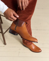 Person wearing brown textured leather ankle boots, adjusting rust-colored corduroy pants while sitting on a chair on a light wood floor.