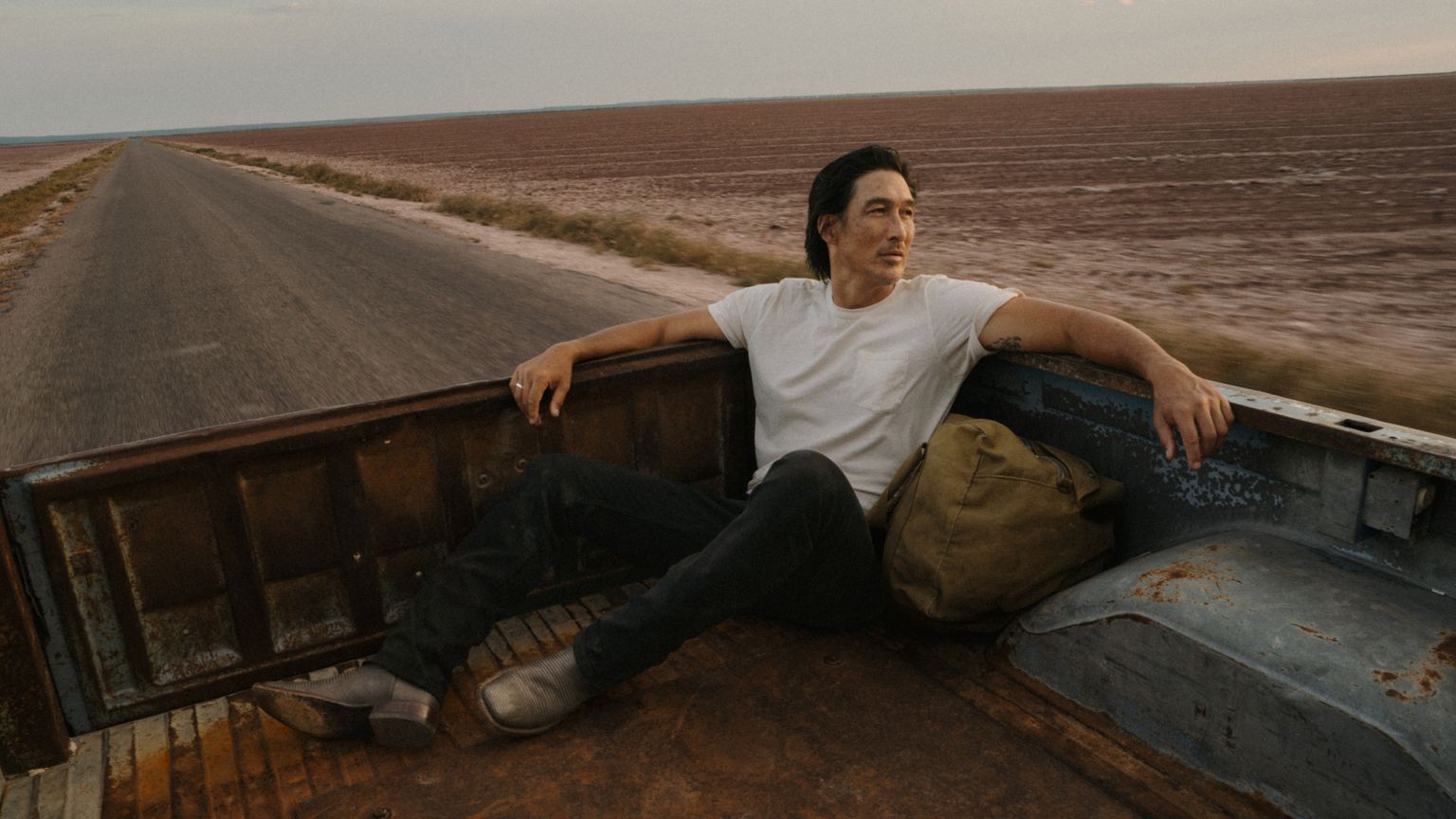 A man in a white t-shirt sits in the bed of a rusty pickup truck with a bag, traveling down an empty road beside a dry, barren landscape.