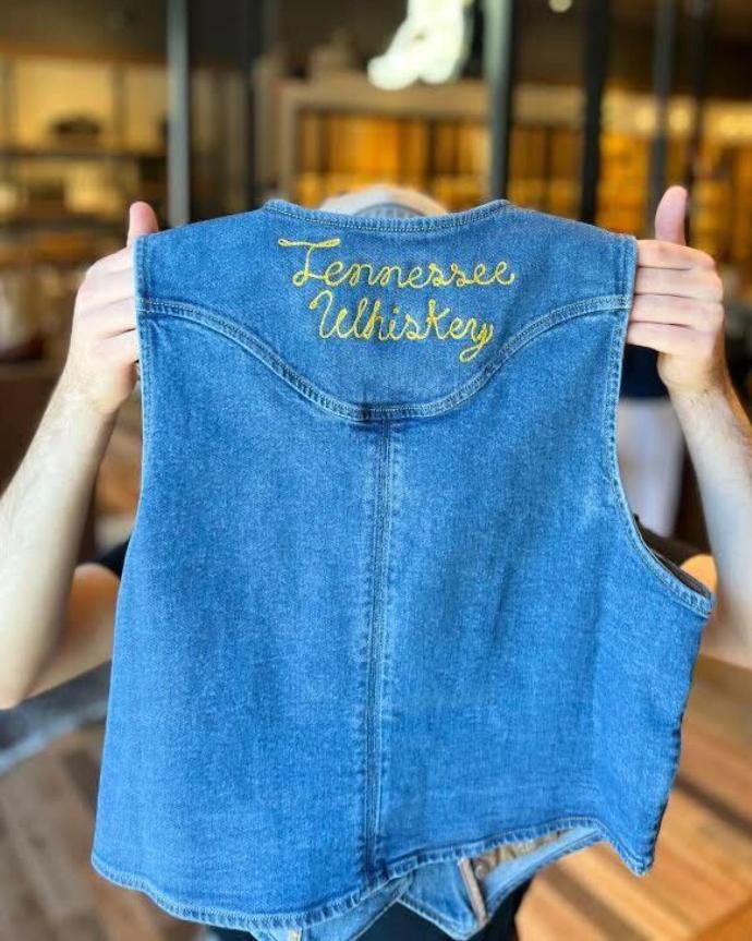 Person holding up a sleeveless blue denim vest with "Tennessee Whiskey" embroidered in yellow on the back.