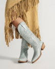 The Annie in Slate Blue Cowhide. Person wearing light blue cowboy boots and a tan fringed suede skirt standing against a plain background.