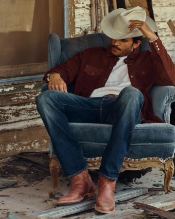 A man in a cowboy hat, brown jacket, jeans, and boots sits relaxed in a worn armchair outside a weathered building, savoring a glass of Scotch as he watches over The Dean Goat nearby.