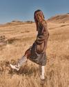 A woman in a patterned dress, brown cardigan, and white boots walks through a dry grassy field under a clear blue sky.
