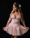 Woman with long, wavy hair wearing a light pink, sleeveless dress with a flared skirt, posing against a black background.