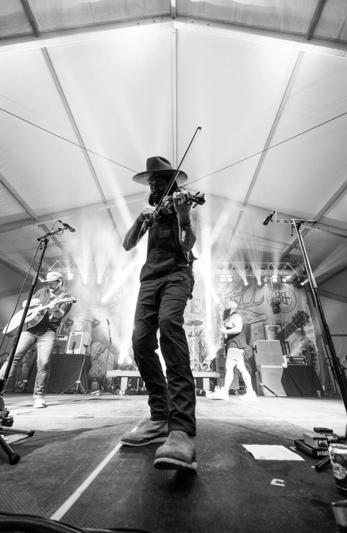 A violinist in a hat performs energetically on stage at a concert, with band members in the background and dramatic stage lighting.