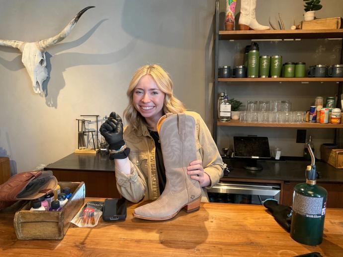 A smiling woman with blonde hair and a tan jacket holds a cowboy boot in a workshop, surrounded by tools and materials. A bull skull is mounted on the wall behind her.