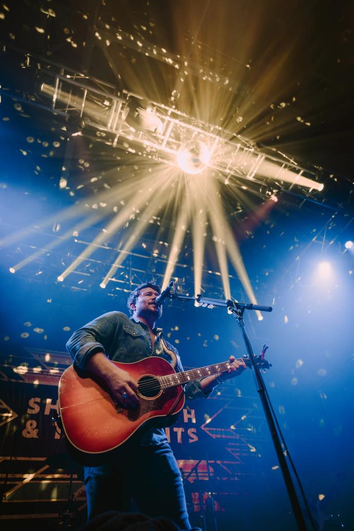 Male musician playing guitar onstage under bright stage lights with a backdrop displaying the text "nash nights".