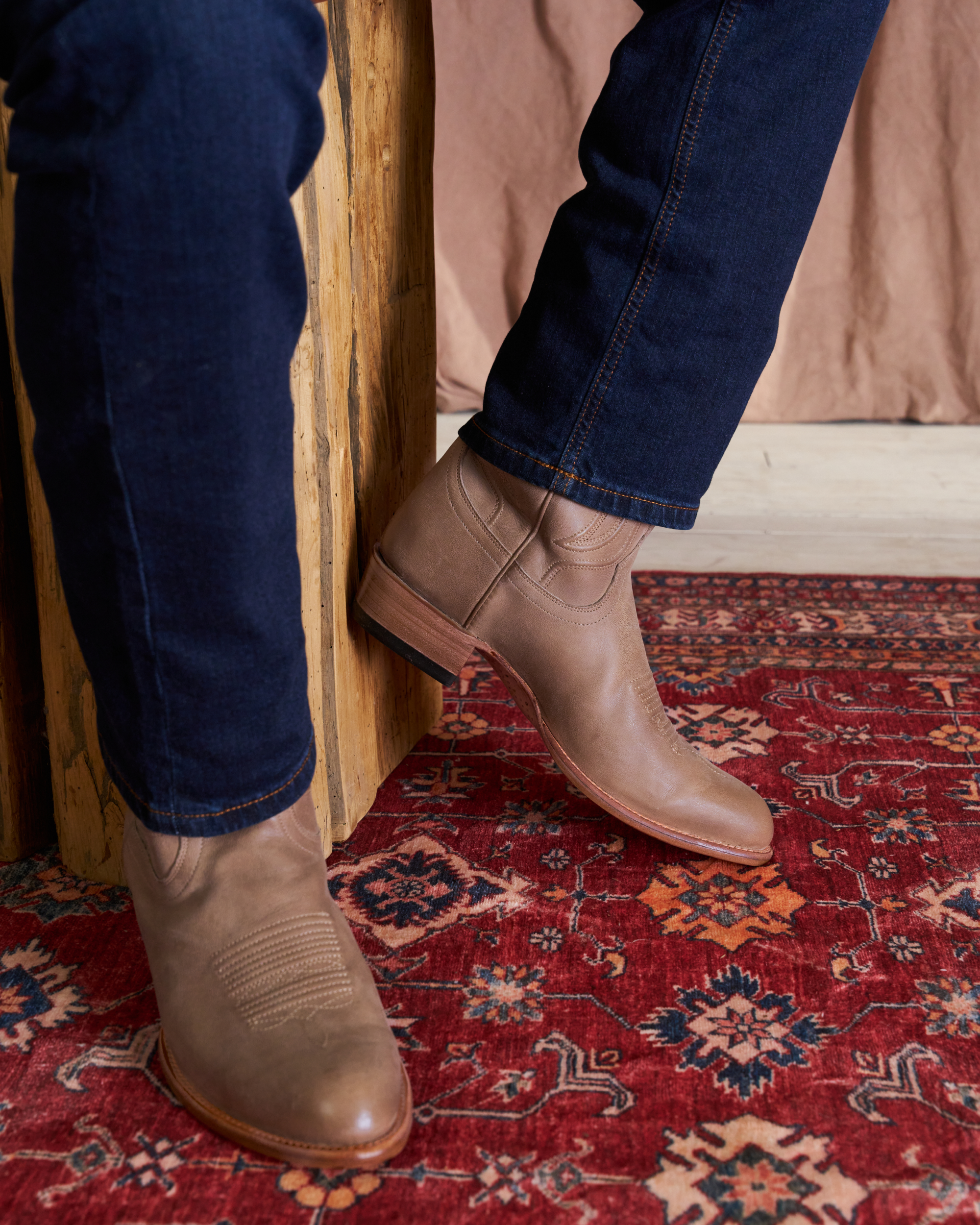A person wearing brown leather boots and blue jeans, standing on a patterned red rug near a wooden structure.