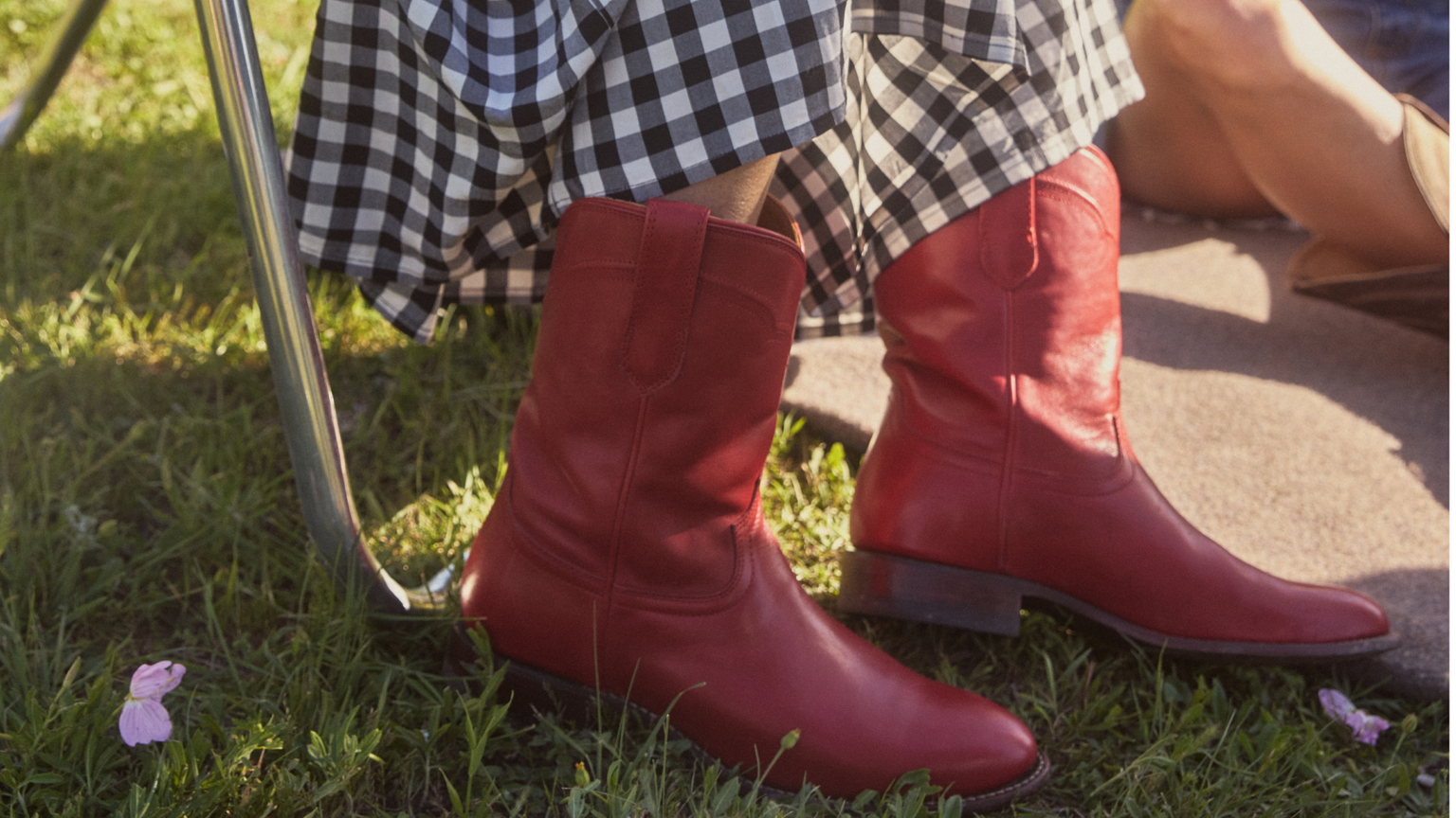 A person wearing red leather boots and a black and white checkered skirt sits outdoors on the grass.