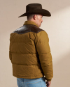 A man wearing a brown cowboy hat, a tan puffer jacket with dark brown shoulder patches, and jeans, stands with his back to the camera in front of a plain background.