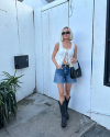 A person wearing a white top, denim shorts, and black boots stands outdoors against a white wall, holding a black bag with a potted plant nearby.