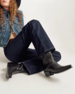 Person sitting cross-legged wearing a denim jacket, dark blue jeans, black cowboy boots, and a black hat against a plain background.