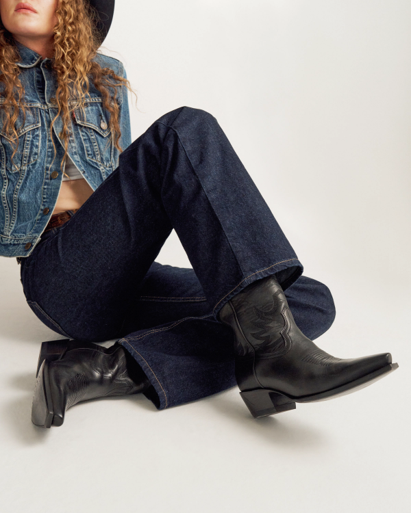 Person sitting cross-legged wearing a denim jacket, dark blue jeans, black cowboy boots, and a black hat against a plain background.